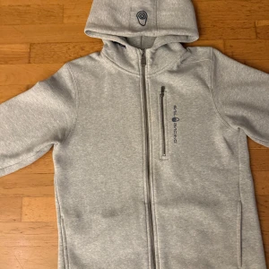 Grå hoodie från Sail Racing - Snygg ljusgrå hoodie från Sail Racing med hel dragkedja och huva. Tröjan har en bröstficka med dragkedja och diskret logga på framsidan. Mjuk bomullsmix och långa ärmar gör den perfekt för kyliga dagar. Stilren och enkel design som funkar till det mesta.