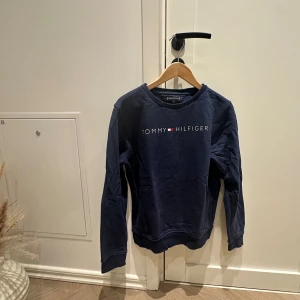 Marinblå sweatshirt Tommy Hilfiger storlek 176 - Snygg marinblå sweatshirt från Tommy Hilfiger med logotyp tryckt över bröstet. Tröjan har rund hals, långa ärmar och mjukt material som är skönt mot huden. Perfekt för dig som gillar klassisk och stilren design.