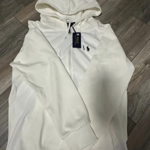 Vit hoodie från Polo Ralph Lauren - Vit hoodie från Polo Ralph Lauren med klassisk logobrodering på bröstet. Tröjan har huva med snörning, dragkedja framtill och ribbade muddar. Materialet är mjuk bomull som känns skönt mot huden. Perfekt för dig som gillar stilrena och enkla plagg.