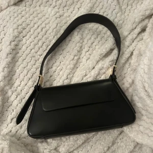 Svart axelväska i skinn med gulddetaljer - Basic svart axelväska i skinn med en geometrisk form och guldfärgade metalldetaljer vid axelremmen. Väskan har ett lock med magnetstängning och ett litet fack inuti. 