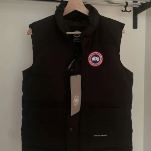 Svart dunväst från Canada Goose - Svart dunväst från Canada Goose, modell Freestyle Crew Vest. Västen har hög krage, två stora fickor framtill och ett broderat rött och vitt märke på bröstet. Materialet är slitstark polyester och dunfyllning för extra värme. Perfekt för kalla vinterdagar.