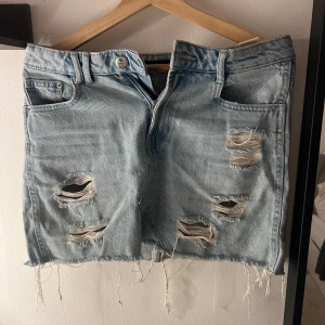 Ljusblå jeanskjol - Säljer en kort ljusblå jeanskjol från Sinsay Denim Collection. Kjolen har slitna detaljer med flera hål och fransig nederkant, klassiska fickor fram och bak samt knapp och dragkedja. Perfekt för dig som gillar en edgy och avslappnad stil.