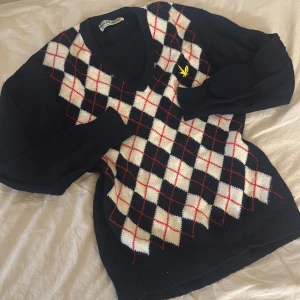 Mörkblå rutig tröja från Lyle & Scott - Snygg mörkblå stickad tröja från Lyle & Scott i 100% lammull. Framsidan har klassiskt argylemönster i vitt, mörkblått och rött samt den ikoniska gula fågeln broderad på bröstet. V-ringad med långa ärmar och ribbade muddar.