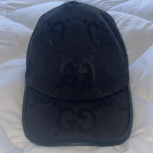 Svart gucci jumbo - Svart keps från Gucci med ikoniskt GG-mönster ton-i-ton över hela kepsen. Klassisk böjd skärm och justerbar rem baktill. Tillverkad i ett slitstarkt tyg med diskret logga på sidan. Perfekt för dig som vill ha en stilren och lyxig accessoar.