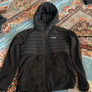 Svart pufferjacka från McKenzie S - Svart pufferjacka från McKenzie med huva och dragkedja framtill. Jackan har quiltade partier upptill och släta ärmar samt två fickor med dragkedja. Perfekt för kyliga dagar och har en sportig vibe.