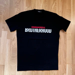 Svart DSQUARED2 Brotherhood t-shirt - Svart t-shirt från DSQUARED2 med trycket 'BROTHERHOOD' i vitt och 'DSQUARED2' i rött på bröstet. Klassisk rund hals och korta ärmar. Perfekt för dig som gillar streetwear och vill sticka ut med en snygg logga.
