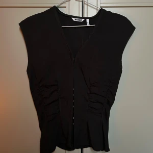 Svart omlottväst från Weekday - Svart väst från Weekday i storlek 36 med snygg v-ringning och omlott-design. Västen har rynkade detaljer längs sidorna som ger en cool siluett. Aldrig använd, prislappen är dock bort klippt🌟 På Bilder ser den ut att vara svart men är mer grå/brun i färgen💕