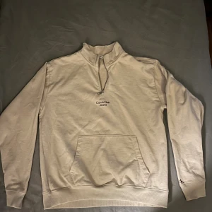 Beige Calvin Klein half zip tröja - Säljer en beige långärmad tröja från Calvin Klein Jeans med half zip och hög krage. Tröjan har en stor magficka och diskret CK-brodyr på ärmen. Perfekt för dig som gillar stilrena och bekväma plagg med enkel design. Helt oanvänd.