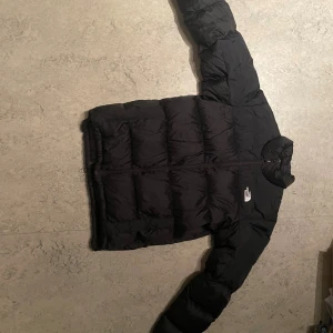 Svart dunjacka från The North Face - Svart dunjacka från The North Face med broderad logga på bröstet och ärmen. Jackan har quiltad design, dragkedja framtill och långärmade ärmar. Perfekt för kalla dagar och har en clean, sportig look som funkar till streetwear. putten liten skada på ena armen.