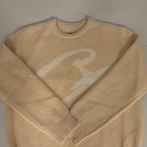 Apricot Corteiz stickad tröja  - Snygg beige stickad tröja från Corteiz med ett stort vitt mönster framtill. Mjuk och skön kvalitet som passar till många outfits. Helt oanvänd i perfekt skick. 