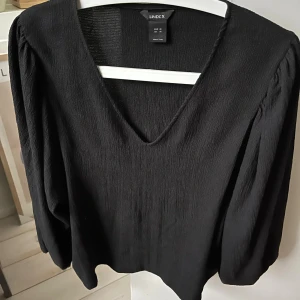 Svart blus med v-ringning från Lindex - Säljer en svart blus från Lindex i storlek M. Blusen har en snygg v-ringad hals och långa ärmar med lätt puff vid axlarna. Materialet är krinklat och ger en cool textur. Perfekt för dig som gillar stilrena och enkla plagg med lite edge.