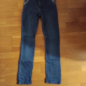 Blå jeans med paljett-detaljer - Snygga blå jeans med raka ben och coola paljett-detaljer på bakfickorna. På ena fickan finns en silverfärgad brosch som ger extra bling. Jeansen har klassisk femficksdesign och är gjorda i mjukt denimtyg. Perfekt för dig som gillar att sticka ut med detaljer. Storlek S 