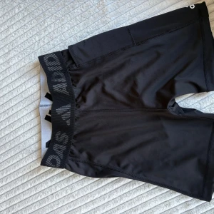 Svarta träningstights-shorts från Adidas - Svarta tighta träningstights-shorts från Adidas med bred resår i midjan där Adidas-loggan syns. Snygg sportig look med diskret logga på benet. Perfekta för gymmet eller löpning, tillverkade i stretchigt syntetmaterial för maximal rörelsefrihet.