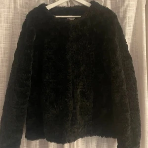 Svart pälsjacka  - Svart pälsjacka med fluffig yta och klassisk rak passform. Jackan har långa ärmar och är helsvart, perfekt för dig som gillar en stilren look. Snygg att matcha med jeans eller kjol.