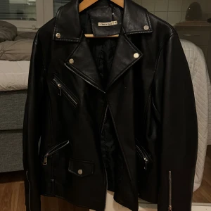 Svart skinnjacka Cherry Koko M - Snygg svart skinnjacka från Cherry Koko i storlek M. Jackan har klassisk bikerstil med flera silvriga dragkedjor, tryckknappar och detaljer på kragen. Perfekt för dig som vill ha en edgy look med coola detaljer och tidlös design. 