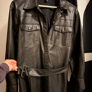 Svart skinnjacka från Soyaconcept - Svart skinnjacka från Soyaconcept med bälte i midjan, stora bröstfickor och tryckknappar framtill. Klänningen har lång ärm och klassisk krage, vilket ger en cool och edgy look. Perfekt för dig som vill sticka ut med en statement piece.