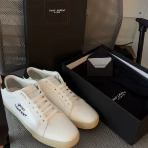 Ysl skor - Snygga vita sneakers från Saint Laurent Paris i skinn med svart detalj på hälen och handskriven logga på sidan. Klassisk rund tå, platt sula och snörning framtill. Perfekta för dig som gillar stilrena och exklusiva skor med lyxig känsla. Storlek 42. Skorna är mycket bra, endast använda ett fåtal gånger!                                                Priset kan diskuteras vid snabb affär!