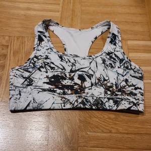 Nike sporttopp med abstrakt mönster - Nike sporttopp i vitt med svart och brunt abstrakt mönster. Toppen har racerback och är ärmlös, med ett snyggt tryckt Nike-logga framtill. Materialet är mjukt och stretchigt polyester, perfekt för träning. Snygg och modern design som sticker ut.