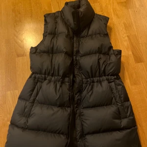 Svart dunväst från Helly Hansen XL - Svart dunväst från Helly Hansen i storlek XL. Västen har hög krage, dragkedja framtill och justerbar midja med snörning. Perfekt för lager-på-lager och håller dig varm med sin quiltade design. Klassisk och stilren look. Nypris 2500 kr