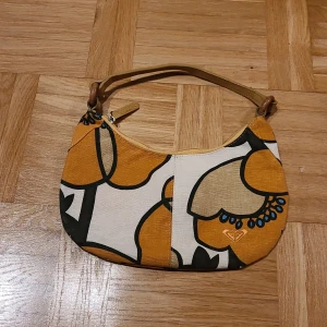 Blommig handväska från Roxy/Quiksilver - Snygg handväska från Roxy med stort blommigt mönster i gult, beige, svart och blått. Väskan har en kort axelrem i skinnimitation och dragkedja upptill. Perfekt för dig som vill sticka ut med färg och mönster.