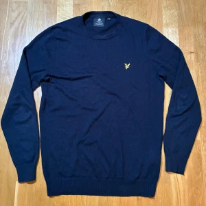 Marinblå stickad tröja Lyle & Scott - Snygg marinblå stickad tröja från Lyle & Scott i storlek M. Tröjan har rund hals, långa ärmar och klassisk gul broderad logga på bröstet. Perfekt för dig som gillar stilrena och tidlösa plagg med en sportig touch. Väldigt bra skick!