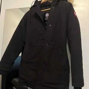 Svart parkas från Canada Goose - Svart parkas från Canada Goose med klassisk röd logga på ärmen och fluffig päls runt huvan. Jackan har långa ärmar, dragkedja och knappar framtill samt två stora fickor. Perfekt för kalla dagar med sin stilrena look och varma material. Den är i väldigt bra skick och har inga skador. Jackan är i storlek XXS