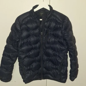 Mörkblå dunjacka Peak Performance - Snygg mörkblå dunjacka från Peak Performance med vågigt quiltad design och hög krage. Jackan har dragkedja framtill och praktiska fickor med dragkedja. Perfekt för kalla dagar och har en clean look som funkar till det mesta.