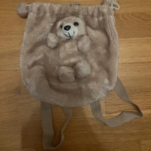 Beige plyschryggsäck med nalle - Beige ryggsäck i mjuk plysch med ett nallehuvud och tassar på framsidan. Väskan har justerbara axelband och snörning upptill.