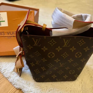 Monogram handväska - Snygg handväska  med klassiskt monogrammönster i brunt och beige. Väskan har en fyrkantig form, beige handtag med röda kanter och en dekorativ rosett samt hänglås i silver. Perfekt för dig som gillar lyxiga detaljer och ikonisk design.