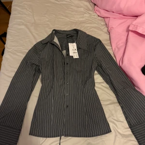 Randig skjorta från Gina Tricot - Snygg skjorta från Gina Tricot i grått med vita smala ränder. Modellen är figursydd med klassisk krage och långa ärmar. Skjortan har knappar framtill och är gjord i mjuk bomull som känns skön mot huden. Perfekt för dig som gillar stilrena och trendiga plagg.