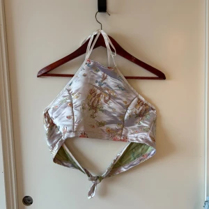 Siden halterneck linnetopp - Unik silvrig linnetopp i siden med halterneck och knyt i ryggen. Toppens yta har broderade drakmotiv i flera färger och insidan är randig i pastellfärger. Perfekt för dig som vill sticka ut med asiatiska vibes och coola detaljer.