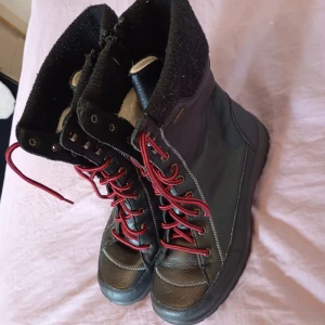 Svarta vinterkängor från Rieker - Svarta kängor från Rieker med röd snörning och grov sula. Utsidan är i skinn och insidan är fodrad med mjuk ull för extra värme. Kängorna har dragkedja på sidan och stickad kant upptill. Perfekta för kalla dagar.