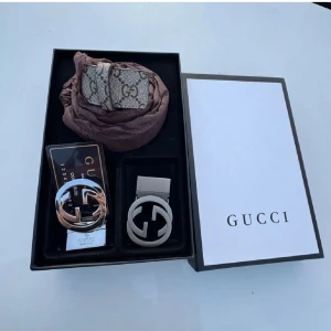 Gucci bälte med GG-logga och box - Säljer ett lyxigt Gucci bälte med ikonisk GG-logga i silverfärgad metall. Bältet har ett klassiskt mönster med GG-print i beige och grå toner, och levereras i originalbox med tillhörande påse. Perfekt accessoar för att lyfta din outfit med exklusiv känsla.