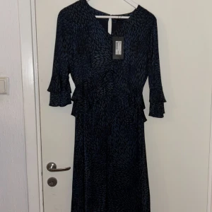 Mörkblå långklänning från Stockh LM - Snygg långklänning från Stockh LM i mörkblått med abstrakt mönster. Klänningen har v-ringning, trekvartsärm med volangdetalj och markerad midja. Perfekt för dig som vill ha en stilren men ändå unik look.