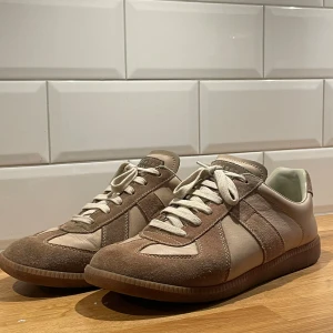 Maison Margiela skor - Maison Margiela sneakers i beige och ljusbrun färg med klassisk rund tå och platt sula. Skorna har snygga paneler i mocka och skinn, samt vita snören. Ikonisk Margiela-etikett på plösen med siffror. Perfekt för dig som gillar stilrena och trendiga sneakers.
