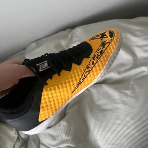 Nike fotbollsskor gul/svart med mönster - Säljer ett par Nike fotbollsskor med grym design i gult och svart, stora mönstrade swoosh-loggor och vita sulor med neon-gula detaljer undertill. Skorna har snörning och är tillverkade i syntetmaterial med prickiga detaljer på ovansidan. Perfekt för dig som vill sticka ut på planen.
