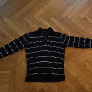 Svart randig polotröja från Selected - Svart polotröja från Selected med vita ränder och tre snygga knappar vid halsen. Tröjan har lång ärm och ribbad krage, perfekt för dig som gillar stilrena detaljer och vill ha en tröja som sticker ut lite extra.