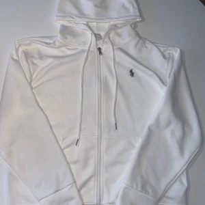 Vit hoodie från Polo Ralph Lauren - Vit hoodie från Polo Ralph Lauren med dragkedja och snörning vid huvan. Klassisk logga broderad på bröstet och två fickor framtill. Mjuk bomullskänsla och långärmad modell som passar perfekt när du vill ha en clean och stilren look.               Priset kan absolut diskuteras!