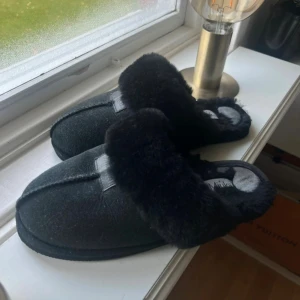 Svarta tofflor från Mjuka med päls - Mysiga svarta tofflor från Mjuka med fluffig päls runt öppningen och mjuk insida. Utsidan är i mocka och har en stilren söm längs mitten. Perfekta för att hålla fötterna varma hemma under kalla dagar.