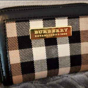 Rutig plånbok från Burberry - Snygg plånbok från Burberry med klassiskt rutigt mönster i svart, beige, vitt och brunt. Plånboken har svarta skinnkanter, guldfärgad metallplatta med Burberry-logga och dragkedja. Insidan har Burberry-tryck och flera fack för kort och mynt.