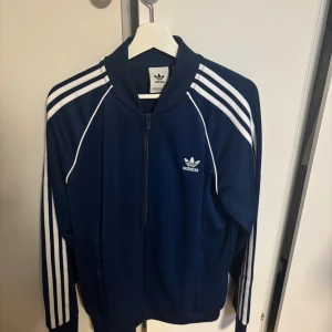 Marinblå Adidas zip-tröja - Säljer en marinblå Adidas zip-tröja med klassiska vita ränder längs ärmarna och Adidas-logga på bröstet. Tröjan har lång ärm, ribbad krage och dragkedja framtill. Perfekt för chill eller sportiga dagar.