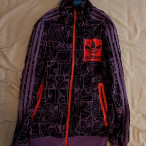 Lila Adidaströja med mönster - Säljer en unik lila tröja från Adidas med grafiskt mönster i svart och lila. Jackan har orange dragkedja, snörning vid kragen och orangea detaljer på fickorna. Stor Adidas-logga i oranget på bröstet. Perfekt för dig som vill sticka ut med streetstyle. Katt finns i hemmet. 