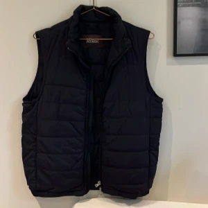 Blå dunväst från Oscar Jacobson - Snygg blå dunväst från Oscar Jacobson med quiltad design och hög krage. Västen har dragkedja framtill och är tillverkad i polyester. Perfekt att slänga över en hoodie för extra värme. Klassisk och stilren look som funkar året runt. Västen är köpt på NK i Göteborg för något år sedan och är sedan dess bokstavligt talat använd 3/4 gånger, den är alltså helt oanvänd och ser likadan ut nu som den gjorde ny i butiken.