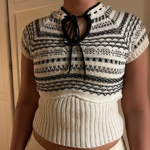 Stickad svartvit topp med knyt - Supersnygg stickad topp i svart och vitt med korta ärmar och coolt grafiskt mönster. Toppen har croppad passform, ribbad midja och knytning vid halsen som ger en unik touch. Perfekt för dig som gillar detaljer och vill sticka ut.