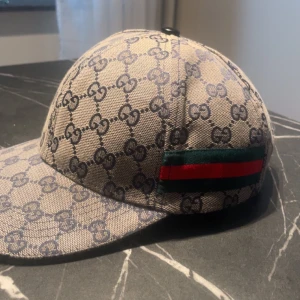Gucci keps med GG-mönster - Säljer en snygg Gucci keps i beige med klassiskt GG-mönster över hela kepsen. På sidorna finns den ikoniska röd-gröna randen. Kepsen är tillverkad i bomullscanvas med justerbar rem bak i svart skinn. Perfekt för dig som vill sticka ut med lyxig streetstyle.
