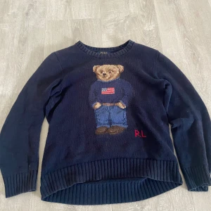 Mörkblå stickad tröja Polo Bear - Mörkblå stickad tröja från Polo Ralph Lauren med ikoniska Polo Bear-motivet framtill. Rund halsringning, ribbade muddar och nederkant. Tröjan har långa ärmar och RL-broderi i rött nedtill. Perfekt för dig som gillar klassisk preppy-stil.