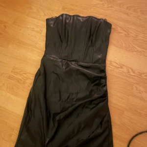 Svart skin klänning från Zara - Säljer en svart axelbandslös klänning från Zara i storlek XS. Klänningen har en snygg, figurnära passform och markerad byst med sömmar. Materialet är glansigt och känns lyxigt, perfekt för dig som vill sticka ut. Supersnygg till klackar och smycken!