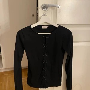 Svart ribbad topp med rosetter från Nelly - Supersnygg svart långärmad topp från Nelly i ribbad bomull. Modellen är figurnära och har rund hals. Tröjan är aldrig använd.