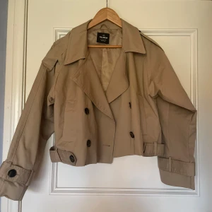 Croppad trenchcoat från Pull&Bear - Slutsåld! Stilren croppad trenchcoat från Pull&Bear i beige bomullsmix. Jackan har dubbelknäppning, bred krage och snygga detaljer med knappar vid ärmslut och axlar. Köptes på Zalando i  juli och har bara använts 4 gånger typ. Helt nytt skick! Kan ta fler bilder om önskat i annat ljus.