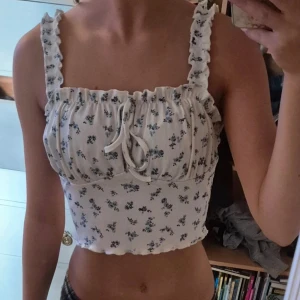 Vit blommig croppad linnetopp - Supersöt croppad linnetopp i vit med blått blommigt mönster från Shein. Toppen har volangkant på axelbanden och en söt knytning framtill. Materialet är mjukt och ribbat, perfekt för sommarens alla dagar.